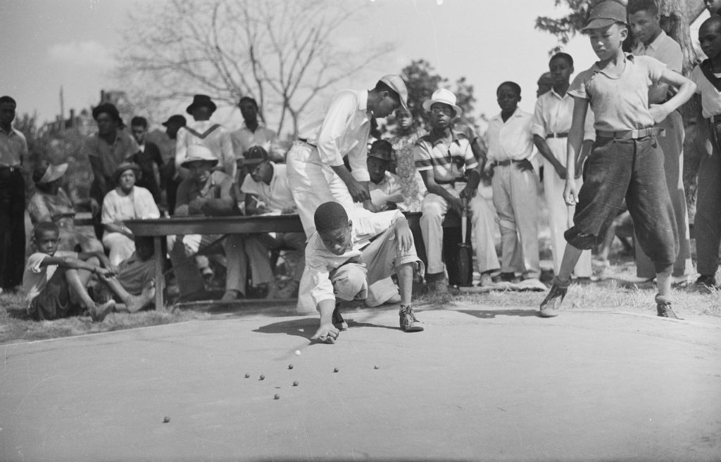 A boy crouches with his knuckle to the pavement, shooting at a scatter of marbles, while a referee bends over him and a crowd of boys and adults watches from benches