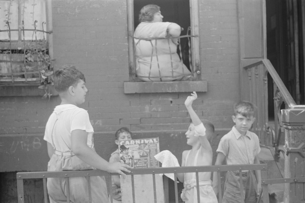Four boys gather at an iron railing outside a brick tenement, one holding up a Carnival game box and another waving, while a woman leans on her arms in the open window above them
