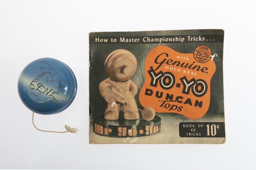 A blue wooden yo-yo engraved Gene beside a Duncan booklet titled How to Master Championship Tricks, Book of 40 Tricks, 10 cents