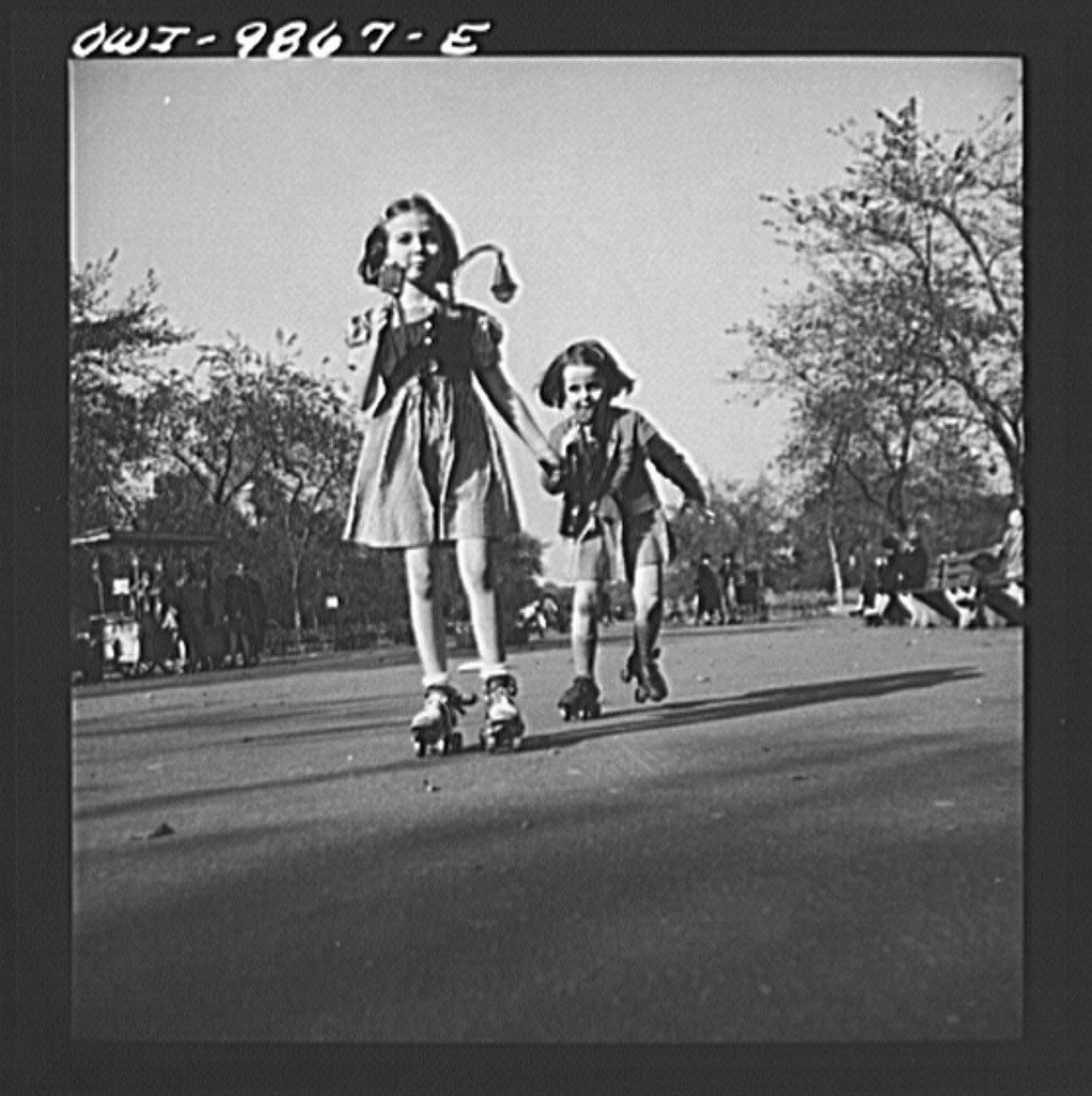 Two smiling young girls in dresses roller-skating toward the camera on a paved path in a park