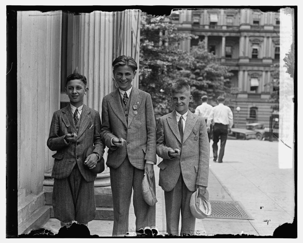 Three teenage boys in jackets and ties standing on a sidewalk beside a columned building, each holding marbles in his hand