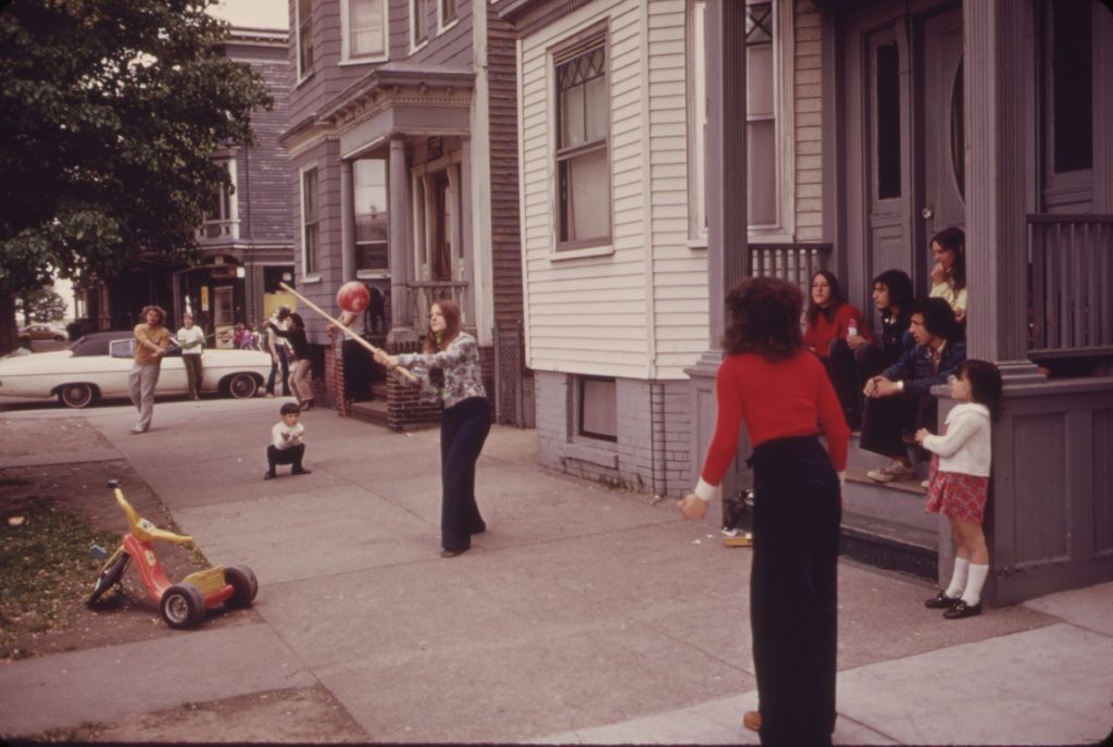 A girl swings a stick at a red rubber ball on a sidewalk lined with wooden houses while another girl waits to field it and older kids watch from a front stoop, a Big Wheel parked on the grass