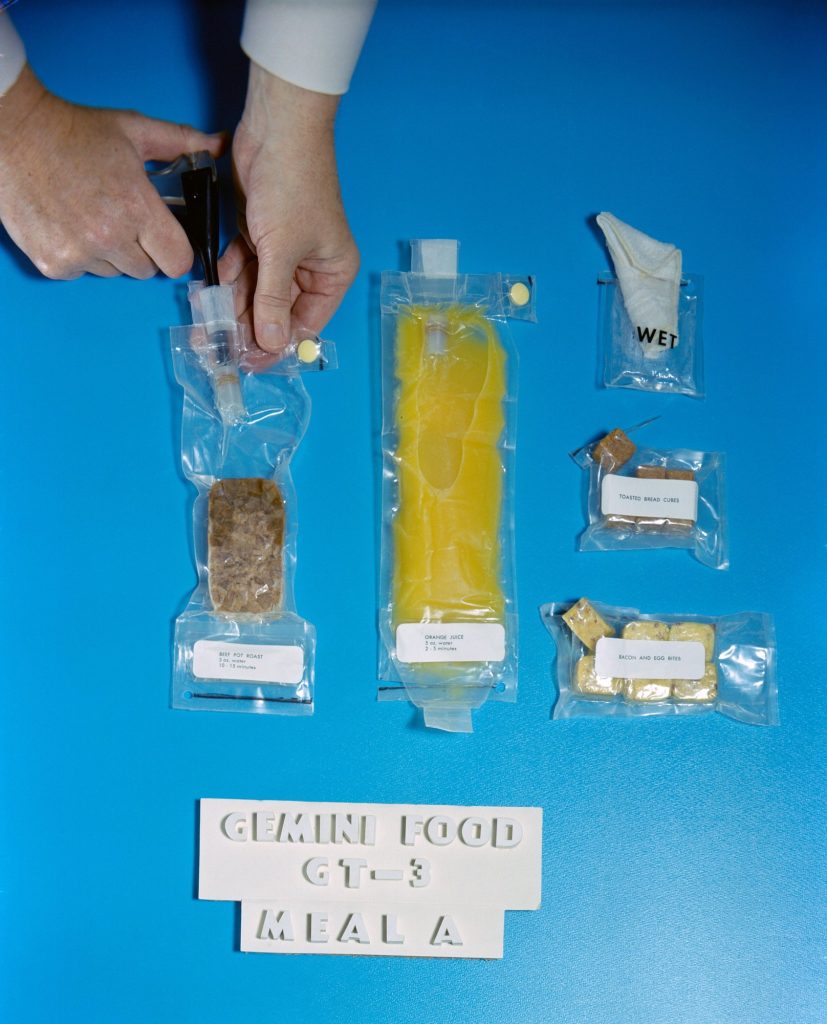 Hands inject water into a clear pouch of dehydrated beef pot roast beside pouches of orange juice, bacon and egg bites, toasted bread cubes and a wet wipe, above a sign reading Gemini Food GT-3 Meal A