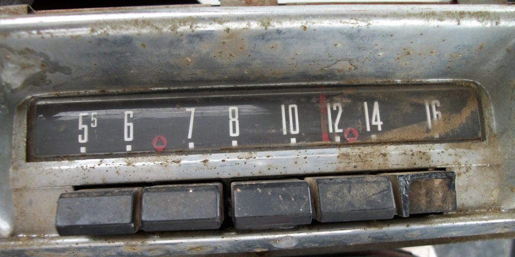 Close-up of a dusty chrome car radio with an AM dial numbered 55 to 16 and small red triangle marks at 6 and 12, above five push buttons