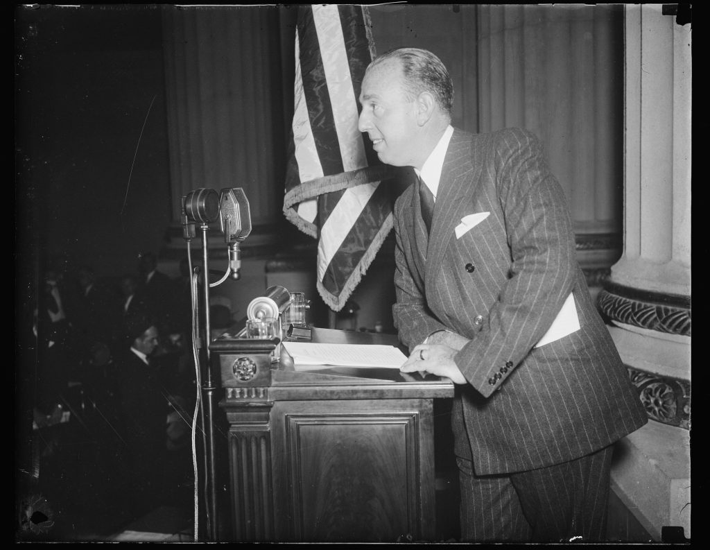 Powel Crosley Jr. in a pinstriped double-breasted suit smiling at a podium with two radio microphones and an American flag behind him