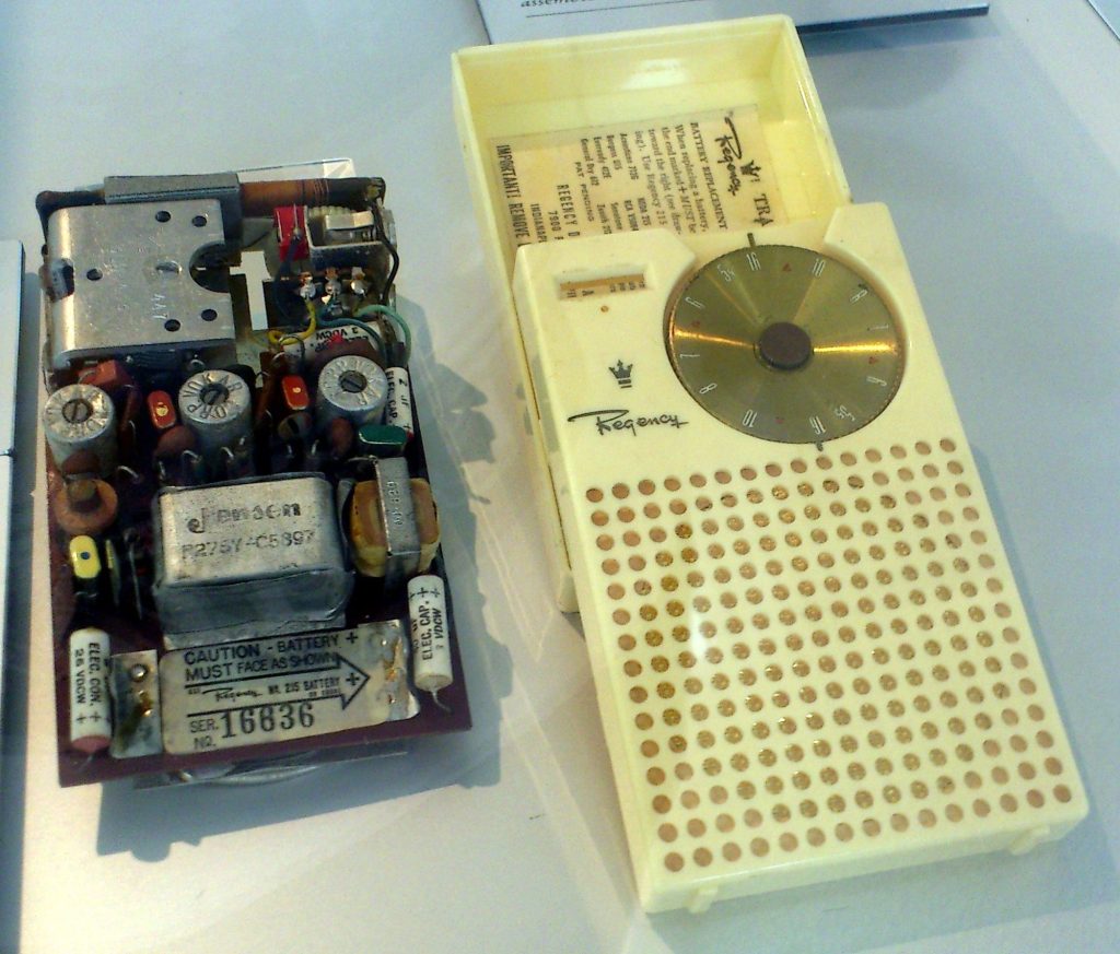 An ivory Regency TR-1 case beside its removed circuit board, crowded with small coils, capacitors and a battery clip labeled Caution, battery must face as shown