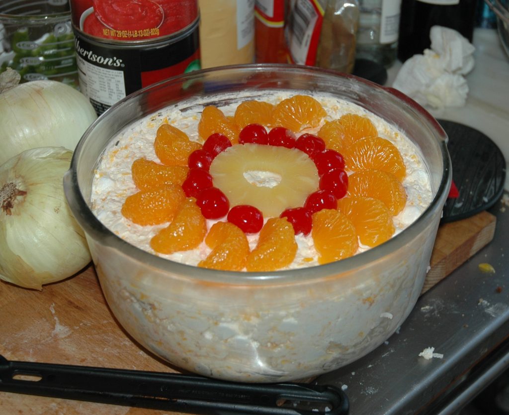 A glass bowl of creamy coconut ambrosia salad topped with a ring of mandarin orange segments, maraschino cherries and a pineapple slice.