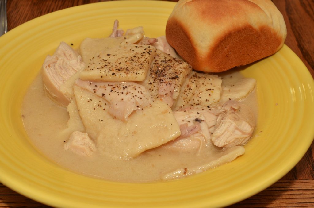 A yellow bowl of chicken and flat rolled dumplings in creamy broth, dusted with black pepper, with a dinner roll on the rim.