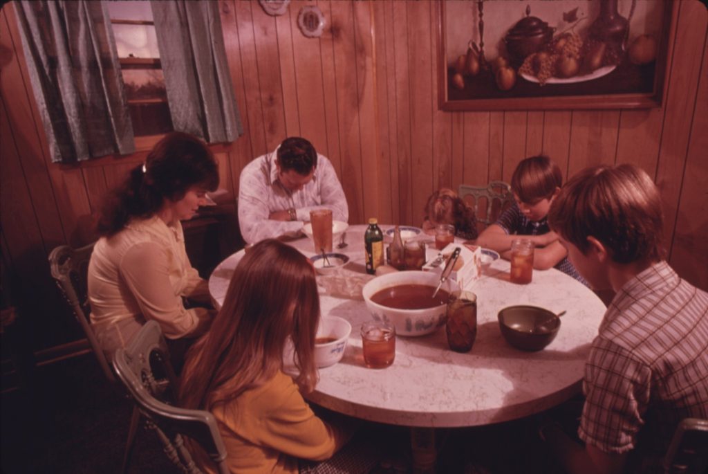 A mother, father and four children bow their heads around a round kitchen table set with bowls of stew, crackers and glasses of iced tea in a wood-paneled kitchen