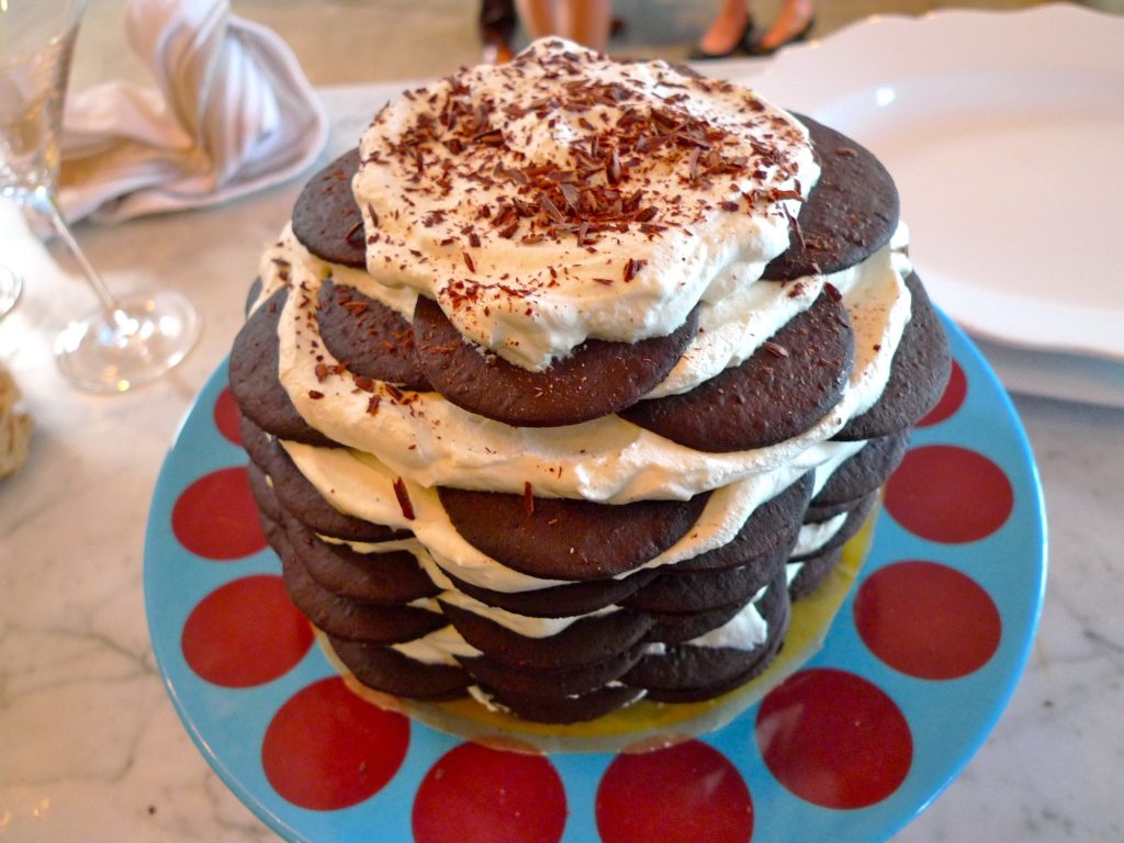 A tall stack of thin chocolate wafers layered with whipped cream and topped with chocolate shavings, on a blue cake stand with red polka dots.