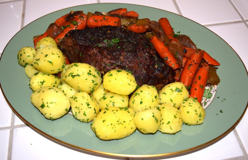 A pot roast on a green oval platter, surrounded by braised carrots and boiled potatoes sprinkled with parsley.