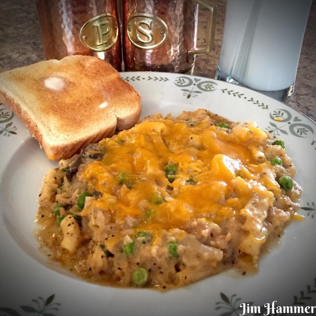 A serving of tuna noodle casserole with peas and melted cheddar on a white plate, next to a slice of buttered toast.