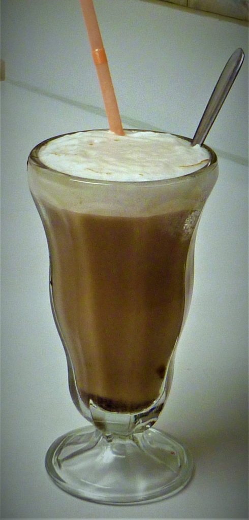 A chocolate egg cream with a thick white foam head in a footed soda-fountain glass, with a pink straw and a long spoon