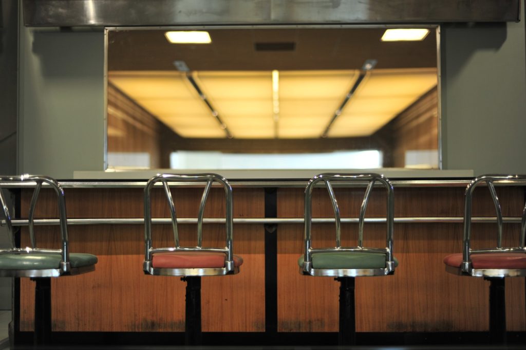 Four chrome stools with green and pink vinyl seats in front of a wood-paneled lunch counter on display in a museum.