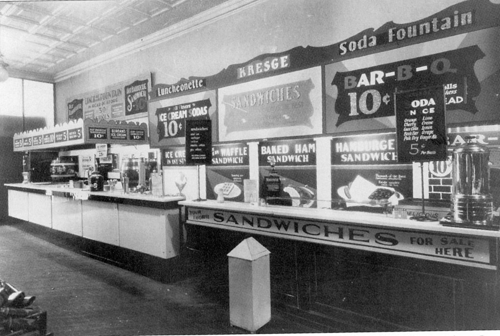 A long store lunch counter under signs reading Kresge Luncheonette and Soda Fountain, advertising 10-cent ice cream sodas, sandwiches, baked ham, hamburgers and 10-cent barbecue, with a coffee urn at the far end.