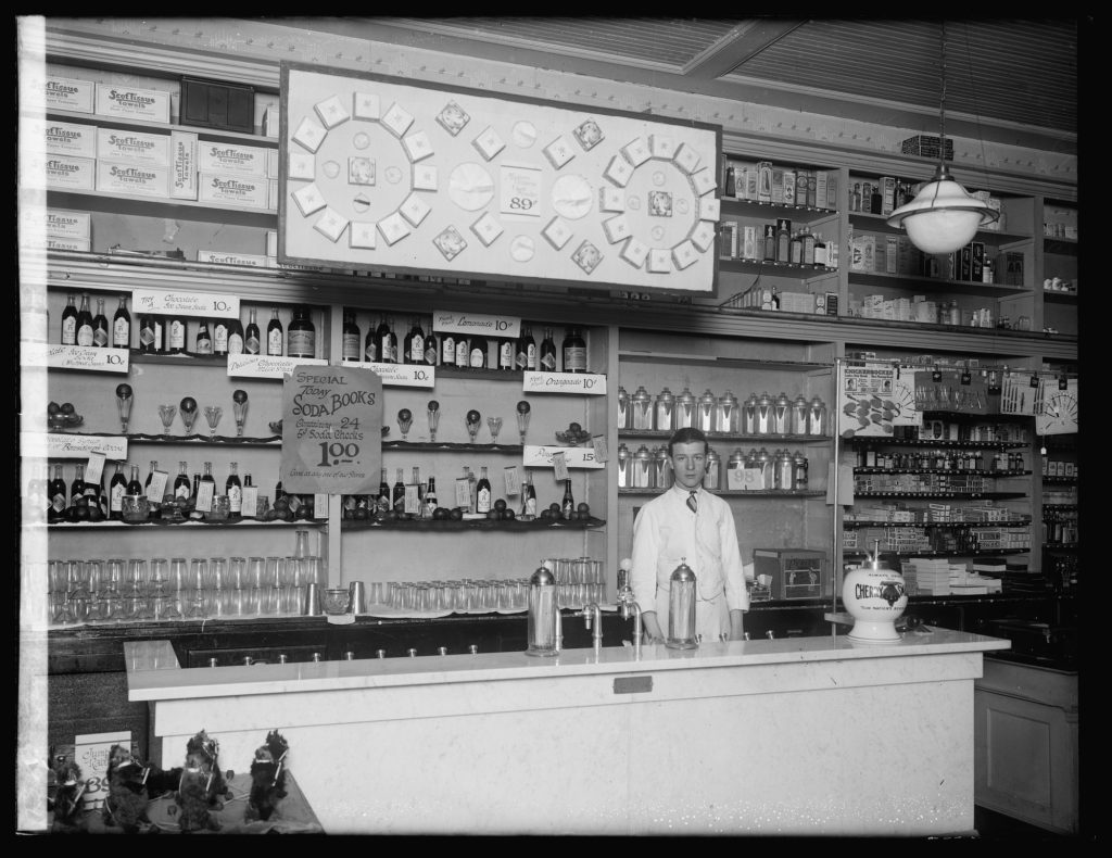 A young clerk in a white jacket stands behind a marble soda fountain, with shelves of syrup bottles and glasses and signs for 10-cent chocolate ice cream sodas, lemonade and orangeade.