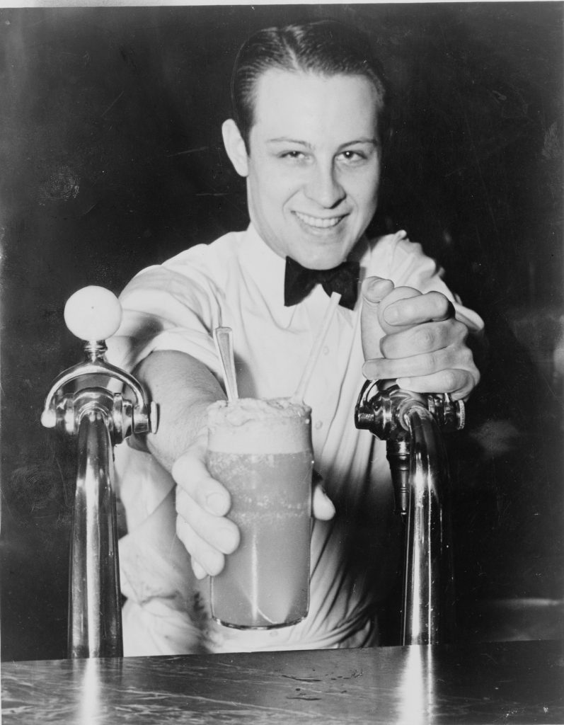 A grinning young soda jerk in a white jacket and bow tie holds out a tall ice cream soda with a spoon and straw between two chrome fountain taps, one hand on a tap handle