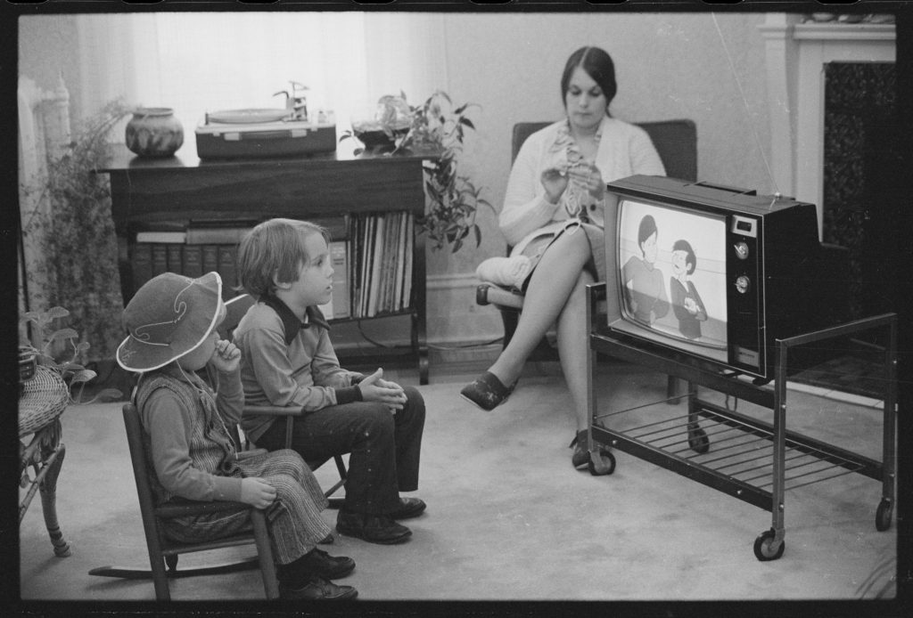 Two young children sit in small chairs watching a cartoon on a portable television on a wheeled cart while their mother knits in an armchair behind them.