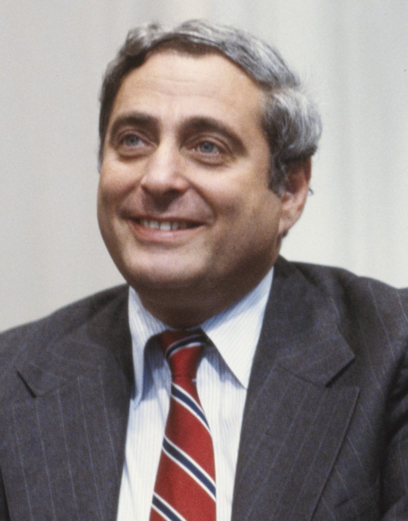 A smiling man with graying hair in a pinstriped suit and red striped tie.