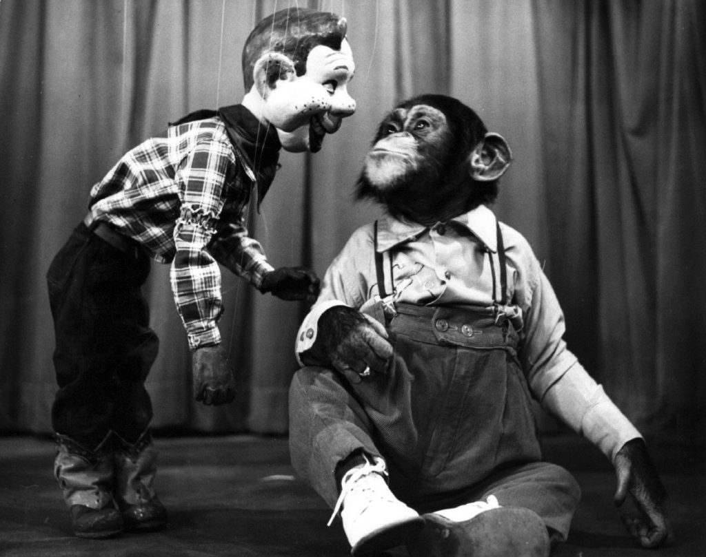 The freckled marionette Howdy Doody in a plaid shirt leans toward a chimpanzee in overalls and sneakers sitting on a stage floor.
