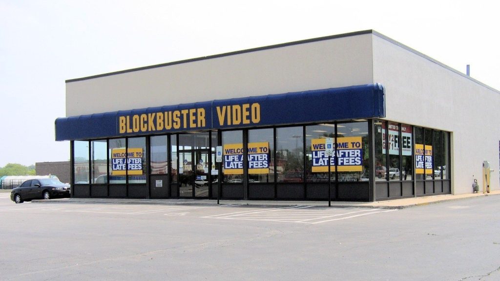 A Blockbuster Video store with a blue awning, its front windows covered with yellow posters reading "Welcome to life after late fees."