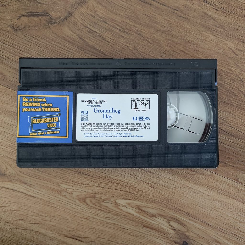 A black VHS cassette of Groundhog Day with a blue and yellow Blockbuster Video sticker reading "Be a friend. REWIND when you reach THE END."
