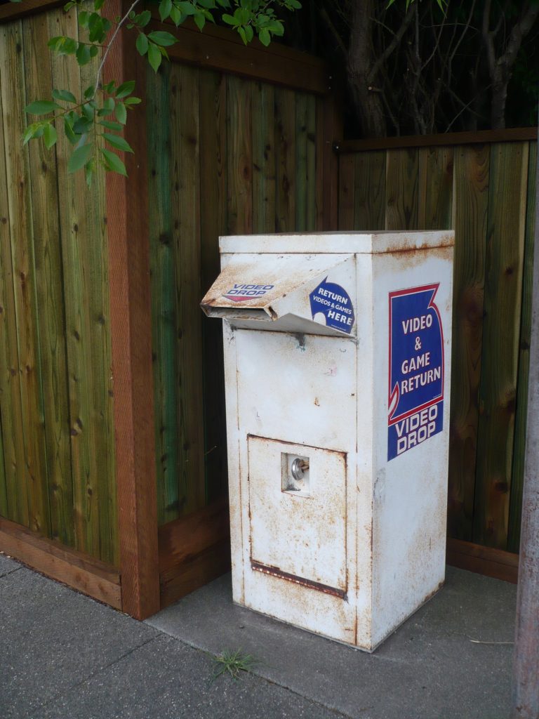 A rust-streaked white metal box on a sidewalk with a slanted slot and stickers reading "Video Drop" and "Video & Game Return."