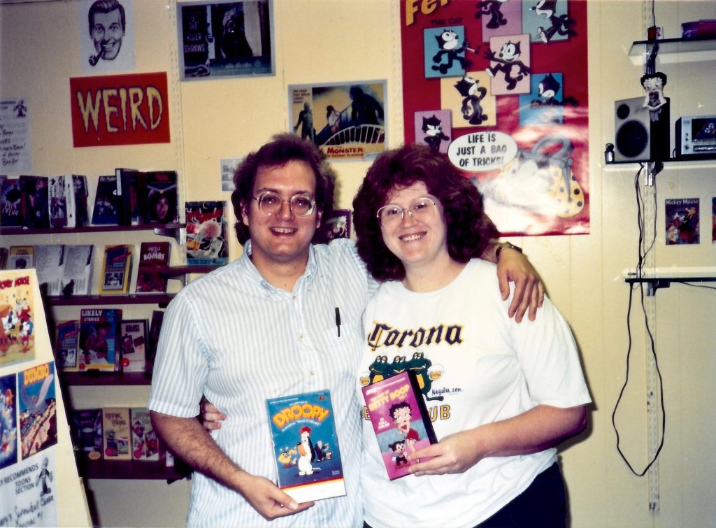 A smiling man and woman in glasses stand arm in arm in a video store, holding a Droopy tape and a Betty Boop tape, below a red sign reading "Weird."
