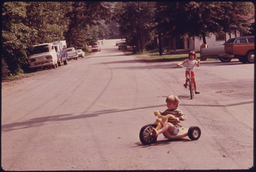 A small child in a striped shirt and plaid shorts rides a yellow low-slung plastic tricycle with a big black front wheel down a paved residential street, while an older child on a bicycle and parked cars and pickup trucks sit behind