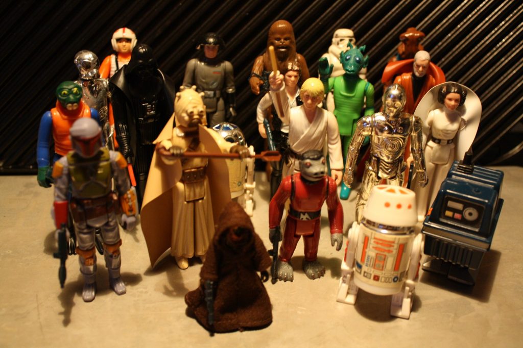About twenty vintage Kenner Star Wars action figures standing in rows on a gray surface, including Darth Vader, Chewbacca, Luke Skywalker, Princess Leia, C-3PO, R2-D2, a Jawa, Greedo and Boba Fett, against a dark striped backdrop