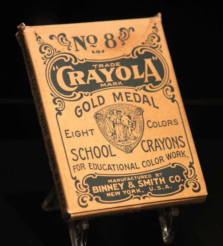 An old tan cardboard box of Crayola crayons on a clear stand, printed No. 8 Crayola Gold Medal Eight Colors School Crayons for Educational Color Work, Binney & Smith Co., New York, U.S.A.