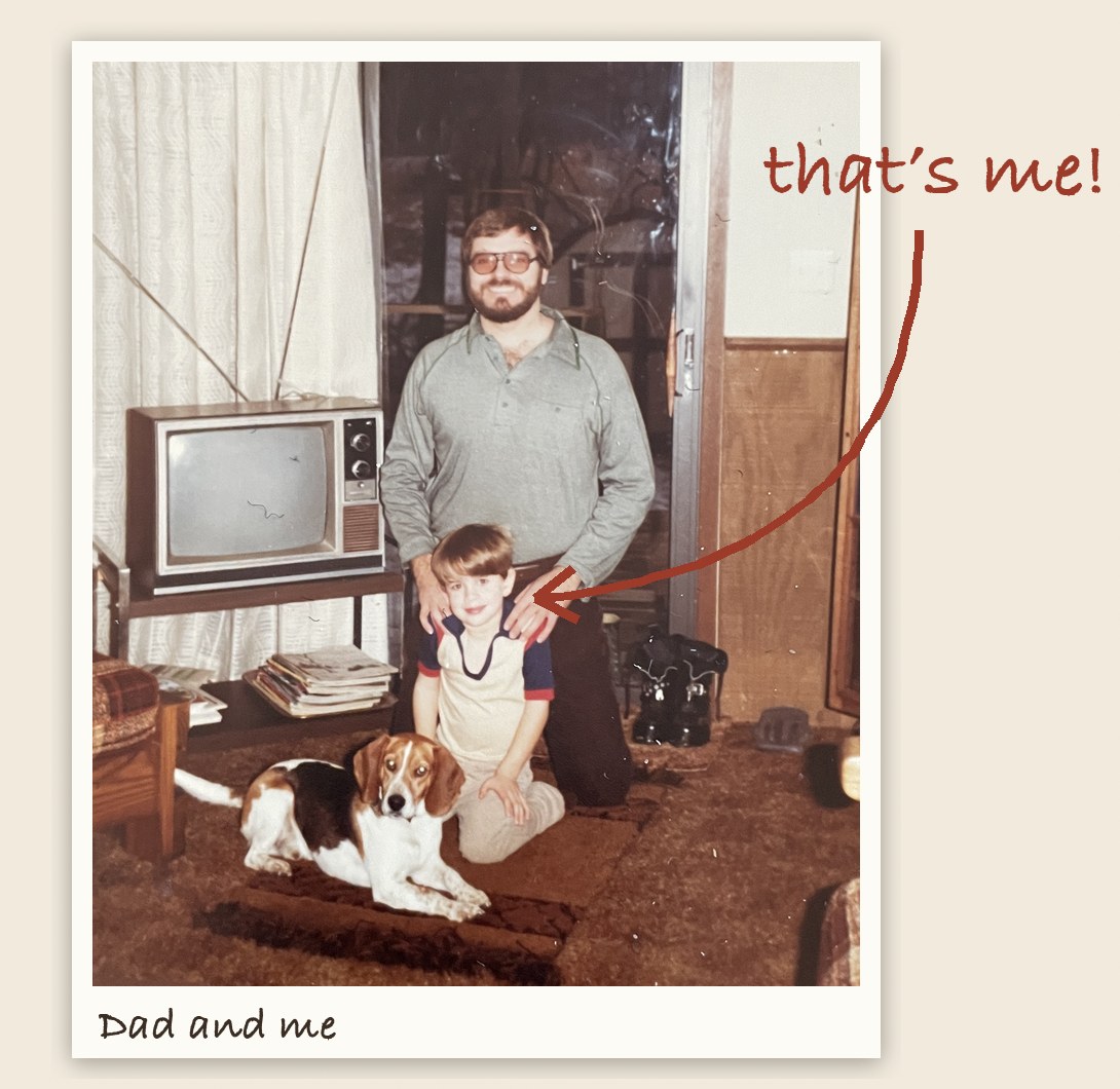 Steve Jones, bearded and in glasses, kneels in a 1970s living room with his young son Cory standing in front of him and a beagle lying beside them, next to a console TV; a handwritten arrow points to Cory with the words “that’s me!” and the print is labeled “Dad and me.”