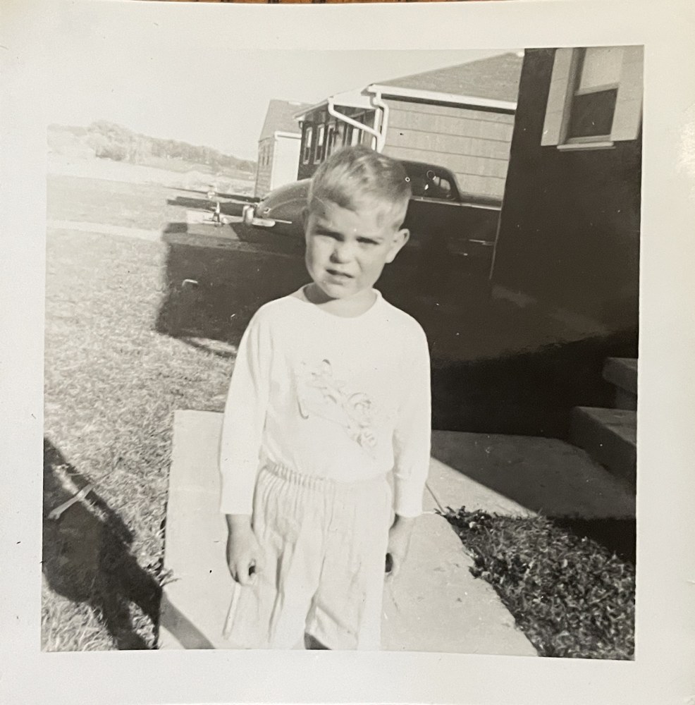 Steve Jones as a little boy in a white long-sleeved shirt, standing on a sidewalk in front of a house and a 1950s car